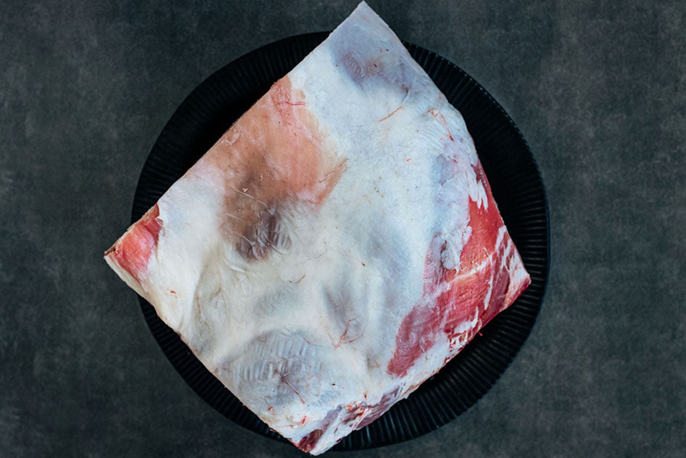 A raw organic lamb shoulder roast with visible fat on it, placed on a black plate.